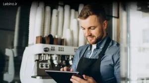 باریستا در حال کار با نرم افزار مدیریت کافی شاپ روی تبلت که یک قهوه ساز در پشت آن قرار دارد که در بالای آن پر از لیوان کاغذی است