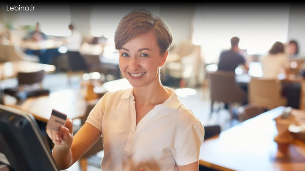 تصویری با کیفیت از یک صندوقدار خندان که در حال استفاده از یک سیستم لمسی و کارتخوان متصل است.سیستم یکپارچه پرداخت رستوران و صندوق فروشگاهی مدرن.